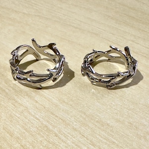 Könnte beinhalten: Zwei silberfarbene Ringe mit einem dornigen Astdesign. Die Ringe liegen auf einer hellbraunen Oberfläche. Die Ringe haben ein offenes, verstellbares Design mit detaillierter, strukturierter Optik.