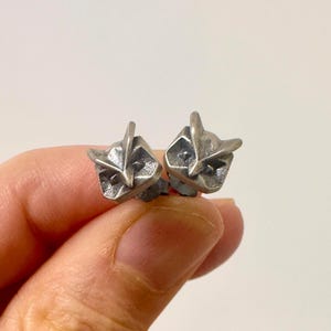 Pode incluir: Um par de pequenos brincos de pino em prata oxidada com formato de cabeças de coruja estilizadas. Cada brinco apresenta um design geométrico texturizado com orelhas pontiagudas, bico triangular e órbitas oculares fundas em acabamento fosco.