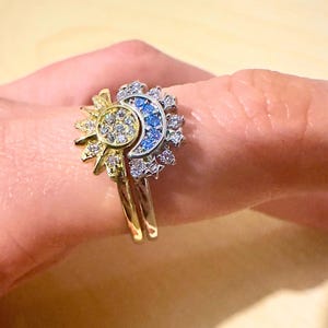 Peut inclure: Une bague en or et argent avec un motif soleil et lune. Le soleil est en or jaune et a une pierre blanche au centre. La lune est en argent et a des pierres bleues autour du bord.