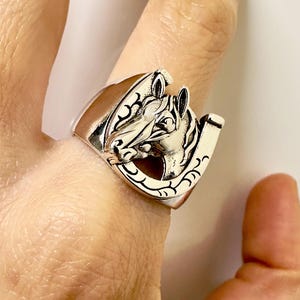 Op de afbeelding: Zilverkleurige ring met een gedetailleerd paardenhoofdontwerp. De ring heeft een hoefijzervorm met decoratief krulwerk. De paardenkop is in profiel, met ingewikkelde details op de manen en het gezicht.