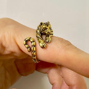 May include: A gold-toned tiger ring. The ring showcases a detailed tiger design with black stripes. The tiger's head and paws are visible, creating a unique piece of jewellery. The ring is wrapped around a finger, adding to its distinctive appearance.