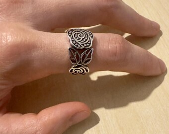 Könnte beinhalten: Ein silberfarbener Ring mit floralem Design. Der Ring hat drei Bereiche: eine Rose oben, ein Paar stilisierter Blätter in der Mitte und eine weitere Rose unten. Der Ring wird an einem Finger getragen.