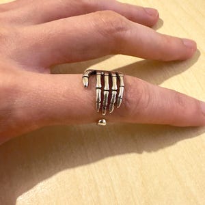 Può includere: Un anello color argento a forma di mano di scheletro. L'anello presenta dettagli di ossa delle dita individuali e viene indossato su un dito. Un accessorio unico e di ispirazione gotica.