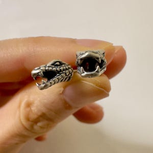 Puede incluir: Dos colgantes plateados con forma de cabeza de serpiente con la boca abierta, revelando dientes. Los colgantes están detallados con escamas y probablemente son para hacer joyas o manualidades. Las cabezas de serpiente miran en diferentes direcciones.