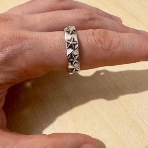 Könnte beinhalten: Ein silberner Ring mit einem sich wiederholenden Sternmuster. Der Ring wird an einem Finger getragen und hat eine strukturierte Oberfläche. Die Sterne sind gleichmäßig um das Band angeordnet.