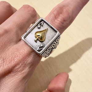 Op de afbeelding: Zilverkleurige ring met het ontwerp van de Aas van Schoppen speelkaart. De ring heeft een rechthoekige voorkant met een gouden schoppensymbool en decoratieve details aan de zijkanten. Een uniek sieraad.