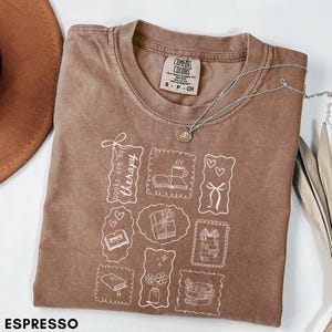 Puede incluir: Camiseta color espresso con un gráfico blanco de libros, café y regalos. La camiseta tiene el texto "books are my therapy" y una etiqueta Comfort Colors. Un collar plateado descansa sobre la camiseta.