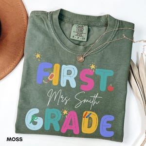 Puede incluir: Una camiseta verde musgo con las palabras "FIRST Mrs. Smith GRADE" en letras de burbujas de colores. La camiseta tiene cuello redondo y mangas cortas. Un collar de oro con un colgante está drapeado sobre la camiseta.