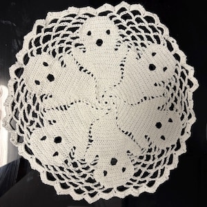 May include: A white, crocheted doily featuring a circular design with six ghost figures. The ghosts have black eye and mouth details. The doily has a scalloped edge and is set against a black background.