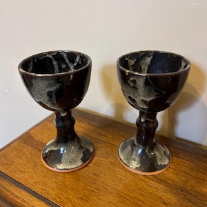 May include: Two black ceramic goblets with white speckles and a brown rim. The goblets have a tall, slender stem and a round bowl.