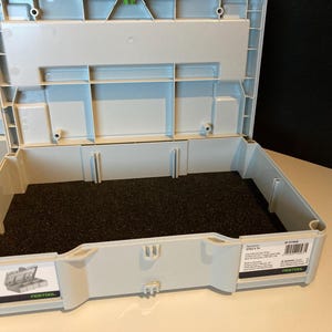 Puede incluir: Una caja de herramientas gris y verde abierta con un inserto de espuma negra. La caja tiene el texto "SYS 1 4" y "Festool" impreso en el lateral.