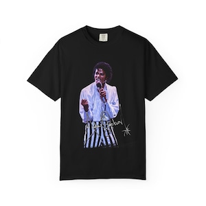 Può includere: T-shirt nero con grafica di Michael Jackson sul palco. L'immagine mostra Jackson con una camicia bianca, pantaloni a righe e un microfono. La sua firma è sotto l'immagine.