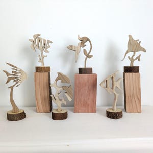 Può includere: Una collezione di sculture in legno raffiguranti vita marina e piante. Le sculture includono pesci, un fiore e una manta, ciascuna montata su una base di legno. I toni naturali del legno creano un'estetica calda e organica.