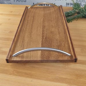 Könnte beinhalten: Ein Holztablett mit zwei Metallgriffen. Das Tablett ist aus hellbraunem Holz mit einer dunkelbraunen Holzverkleidung. Die Griffe sind silberfarben und gebogen.