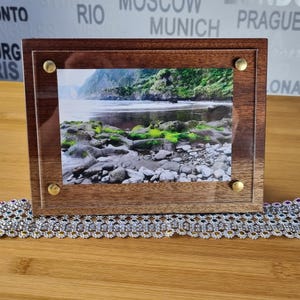 Könnte beinhalten: Ein Holzbildrahmen mit einer klaren Acrylglasscheibe und Messingakzenten. Der Rahmen enthält ein Foto eines felsigen Strandes mit grünem Moos und einem Gewässer im Hintergrund.