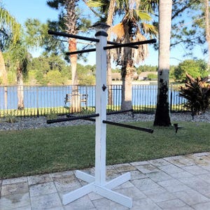 May include: A white wooden towel rack with black horizontal bars for hanging towels. The rack has a decorative top and a cross-shaped base. It is outdoors on a patio with a lake and palm trees in the background.