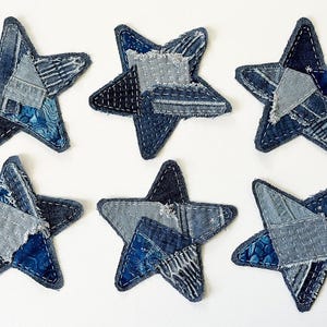 May include: Six star-shaped fabric patches crafted from various shades of blue denim and cotton textiles. Each star features a patchwork design with visible white stitching, frayed edges, and a mix of light, medium, and dark blue fabric textures.