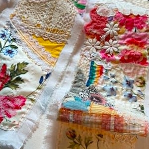 May include: A textile collage featuring various fabric scraps, including floral prints, white lace, yellow fabric, and a small rainbow patch. The pieces are joined with visible stitching, incorporating embroidered flowers, pink fabric, and white daisy appliqués.