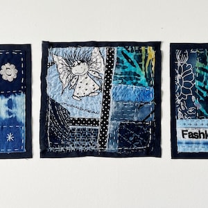 May include: Three square fabric collages in shades of blue, featuring patchwork patterns, white stitching, and embroidered details. The left piece displays the text EXTRA!, the center piece features a small angel illustration, and the right piece shows the word Fashion.