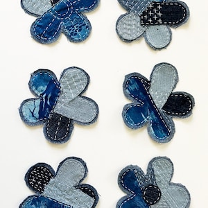 May include: Six flower-shaped fabric patches made from various shades of blue denim and cotton textiles. Each patch features visible white hand-stitched embroidery, including cross-stitch and running stitch patterns, with frayed edges on the petals.