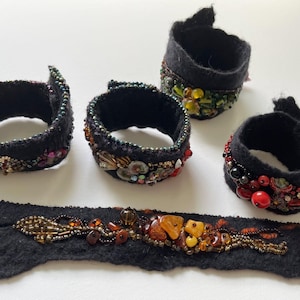 May include: Four black felt cuffs featuring intricate beadwork and one flat black felt strip adorned with amber-colored stones and metallic beads. The cuffs are circular with visible stitching and varied embellishments in red, green, and gold tones.