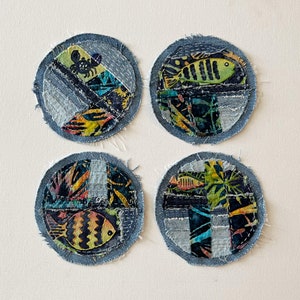 May include: Four round denim coasters with colorful patchwork designs featuring fish, a crab, and floral patterns. The coasters are made from repurposed denim fabric and have a frayed edge.