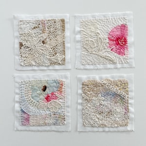 May include: Four square fabric art pieces with intricate lace and floral designs. Each piece features a different arrangement of textures and colors, including cream, pink, and blue, set against a white background. The art pieces are likely handmade.