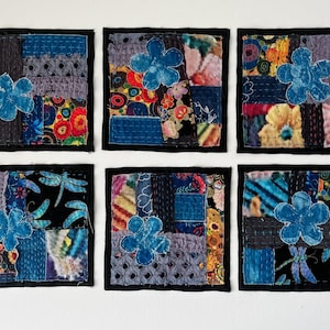 May include: Six square fabric coasters with a patchwork design. Each coaster features a blue flower applique and a variety of patterned fabrics in shades of blue, purple, and floral prints. The coasters are outlined with black stitching.
