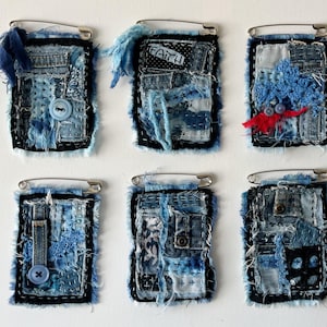 May include: Six handmade denim brooches with safety pin closures. Each brooch features a unique patchwork design in shades of blue, with frayed edges and decorative stitching. Some include buttons and fabric embellishments.
