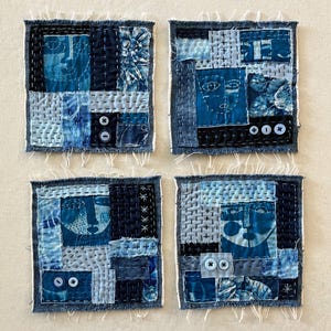 May include: Four square textile art pieces with abstract faces, crafted from denim and various blue fabrics. Each piece features hand-stitched details, buttons, and frayed edges, creating a unique, textured appearance. The artwork is arranged on a neutral background.