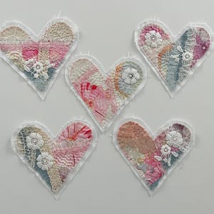 May include: Five handmade heart-shaped fabric ornaments. Each heart features a patchwork design with lace, floral embellishments, and a raw edge. The color palette includes pinks, blues, and creams, creating a soft, vintage aesthetic.