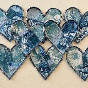 May include: Nine blue denim hearts with a patchwork design. Each heart is made of different pieces of denim fabric, sewn together with white thread. The hearts are arranged in a cluster, with some overlapping.