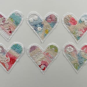 May include: Six handmade fabric heart ornaments. Each heart features a patchwork design with floral and lace accents. The color palette includes pink, blue, and white, with decorative stitching around the edges.