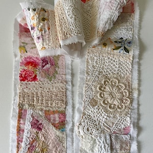 May include: A textile collage featuring layered fabric scraps, intricate white lace, and floral-printed cotton patches in shades of pink, cream, and white. The composition includes frayed edges, textured embroidery, and various lace patterns stitched together.