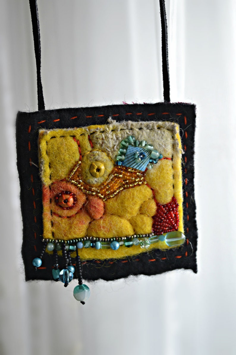 May include: A yellow, orange and blue felt pendant with beads and a black cord. The pendant is a square shape with a stitched border.