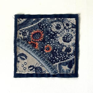 May include: A square fabric patch featuring a dark blue and beige patchwork design with visible stitching. The textile displays embroidered orange sun-like motifs, white dotted patterns, and a dark blue crab graphic on a light beige background.