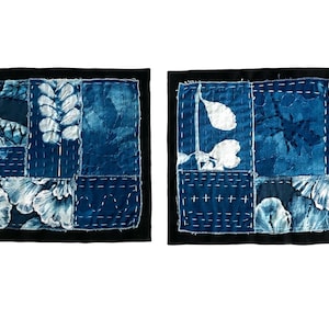 May include: Two square fabric panels featuring indigo blue patchwork with white botanical prints and visible white hand-stitched running thread patterns. Each panel is framed by a solid black border with frayed edges and a quilted texture.