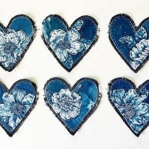 May include: Six heart-shaped fabric patches featuring a deep blue denim base with frayed edges. Each patch displays a detailed white floral illustration with intricate petals and leaves, showcasing a consistent botanical design across all pieces.