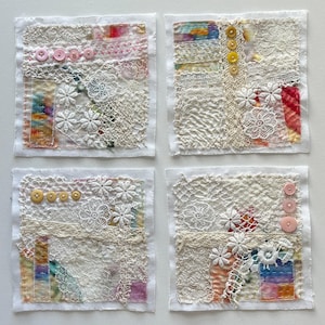 May include: Four square textile art pieces. Each features a white fabric base with layered textures of lace, embroidery, and colorful fabric scraps. Decorative buttons and floral lace accents add detail. The pieces are arranged in a grid.