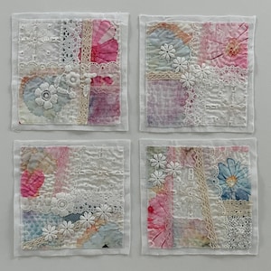May include: Four square fabric art pieces with a patchwork design. Each piece features floral patterns, lace, and embroidered details in shades of pink, blue, and white. The squares are bordered with white fabric.