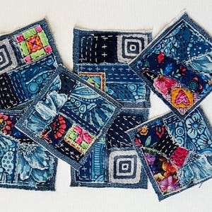 May include: A set of five square fabric coasters featuring a patchwork design of blue denim and various floral, geometric, and abstract patterned textiles. Each coaster is finished with visible white stitching and frayed edges in a collage style.