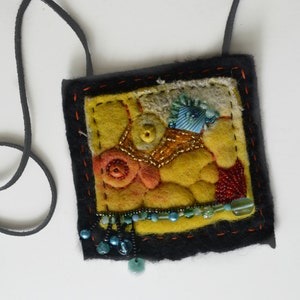 May include: A yellow felt pendant with a black leather cord. The pendant is decorated with colourful beads, buttons, and fabric scraps.