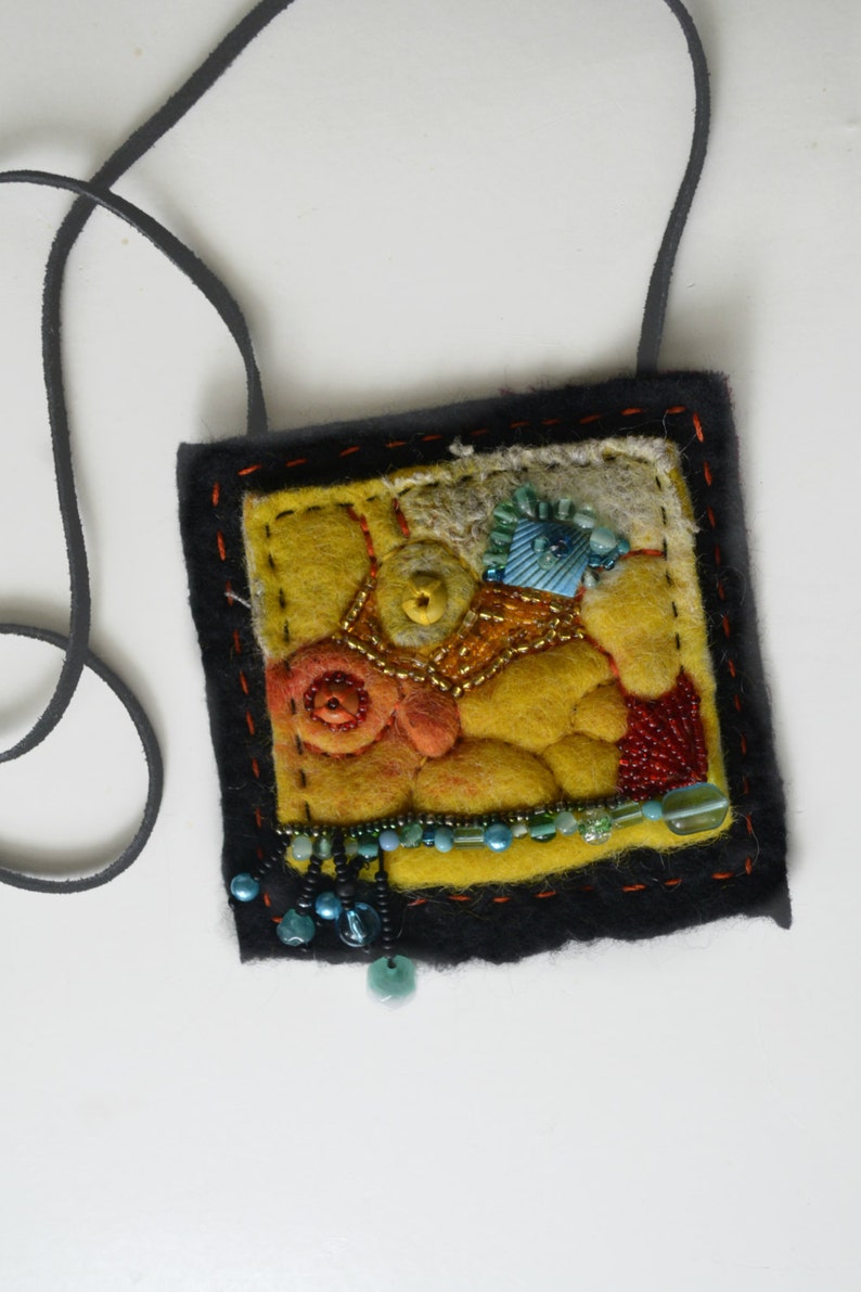 May include: A yellow felt pendant with a black leather cord. The pendant is decorated with colourful beads, buttons, and fabric scraps.