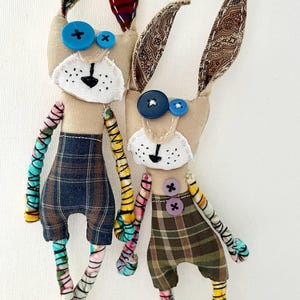 May include: Two handmade fabric rabbit dolls with long ears, button eyes, and stitched faces. The dolls wear plaid shorts and have multi-colored, patterned fabric arms and legs. One rabbit has red plaid ears, while the other has brown paisley ears.