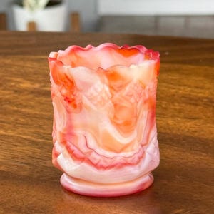 May include: A small, decorative glass vase with a ruffled rim. The vase features a blend of orange, red, and white colors, with a swirled pattern. The exterior has a textured design. The vase is approximately 3 inches tall.