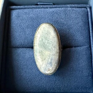 May include: A ring featuring a large, oval-shaped, pale greyish-green stone with a smooth, polished surface. The stone is set within a simple, thin metal bezel that follows the oval contour, highlighting the natural variations and luster of the gem.