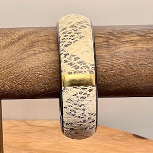 May include: A white and black bangle bracelet with a gold accent. The bracelet has a snakeskin pattern.