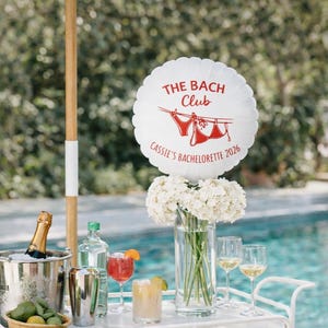 May include: A white cart with drinks, glasses, and a floral arrangement sits by a pool. A round white balloon with the text "THE BACH Club" and "Cassie's Bachelorette 2026" is displayed. An umbrella with green and white stripes provides shade.