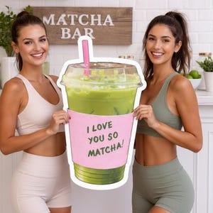 May include: Two women holding a large, cut-out image of a matcha drink in a clear plastic cup with a pink sleeve that reads "I LOVE YOU SO MATCHA!". The drink is green, and the straw is pink. A wooden sign that says "MATCHA BAR" is in the background.