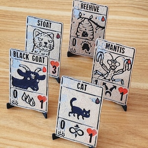 May include: Five illustrated game cards with black stands. The cards feature animal illustrations and text, including "Stoat," "Beehive," "Black Goat," "Mantis," and "Cat." Each card has a unique design with symbols and numbers, set against a beige background.